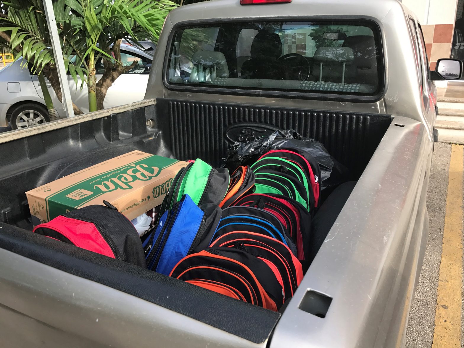 A dozen backpack in variety of bright colors sit in the bed of tan pick up truck along side a large cardboard box.
