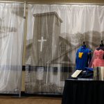A few items from a mobile exhibit. Tall silk curtain with images of a church and other landscapes create the backdrop. Off to the left a table is set up to display collection items such as year books, graduation cap. Next to the table are dress forms displaying a pink strapless prom dress and a blue and gold varsity letter sweatshirt.