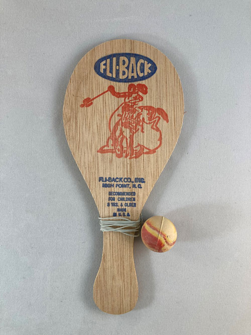 Paddle-shaped board with an image of a cowboy or gaucho on horseback swinging a rope with a ball on the end. The word "Fli-Back" is printed above the image.