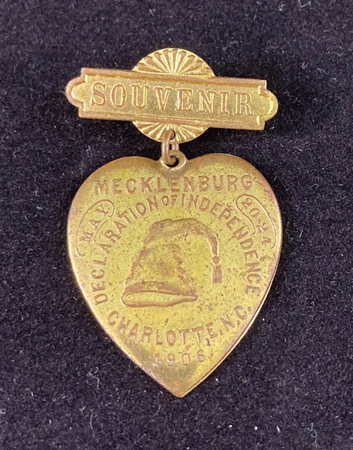 Gold-colored, heart-shaped metal with the words Mecklenburg Declaration of Independence, Charlotte, N.C. and an image of a night cap