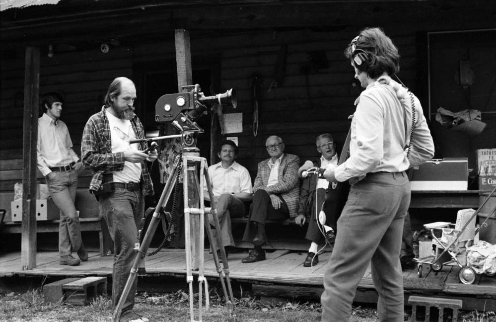 Filmmaker Les Blank and sound person, Mike Seeger, standing in front of Tommy Jarrell's home. Tommy Jarrell and others are seen sitting on the porch.