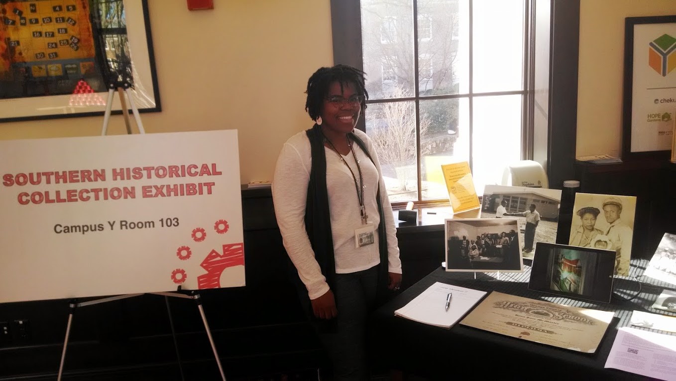 Southern Historical Collection staff member Chaitra Powell next to a table with archival records, including images related to HBSTA history, and a sign reading, "Southern Historical Collection Exhibit." 