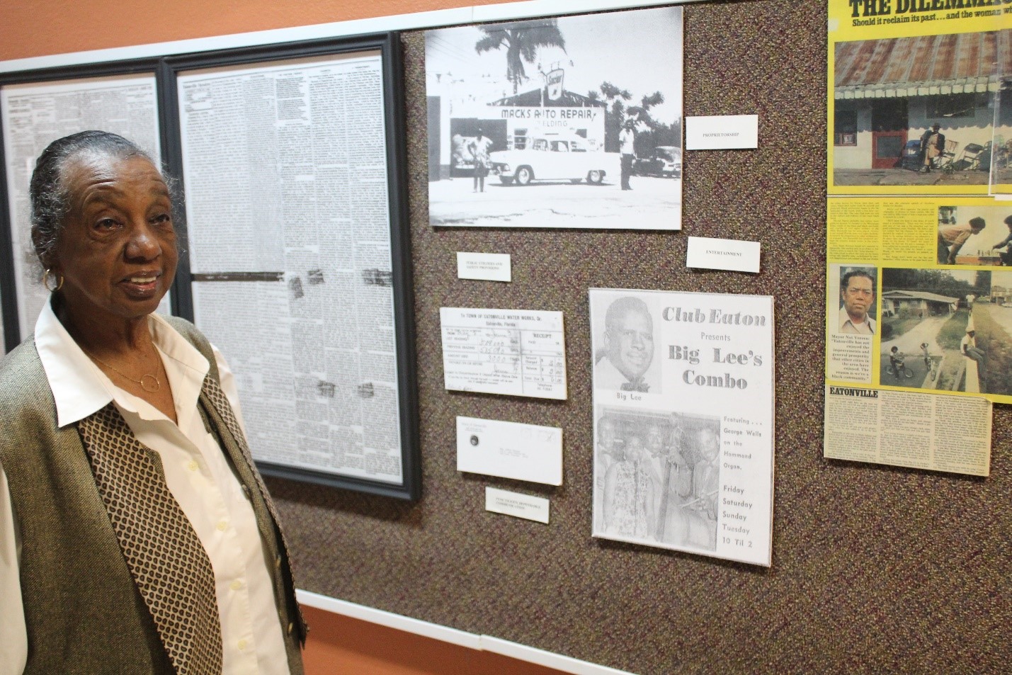 Mrs. Maye St. Julien next to a bulletin board with historical images and articles displayed with captioned labels next to each.