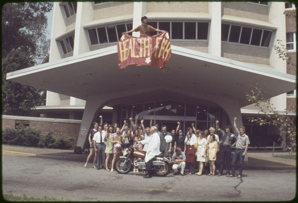 Majority white group of students, many smiling, with their fists raised in the air. An older person is in front of the group wearing a white lab coat on a motorcycle.