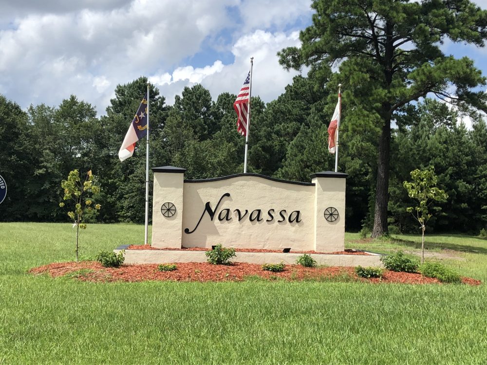 Photograph of the Navassa town sign. Three flags, the North Carolina flag, American flag, and a third flag, stand behind the town sign. 