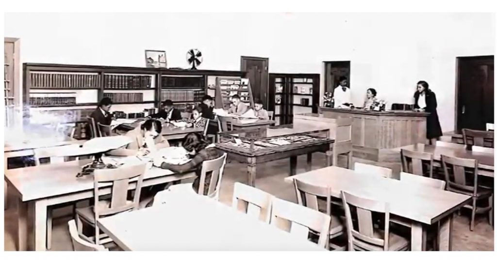 Library room with book shelves and tables and chairs with seated patrons and staff at the front