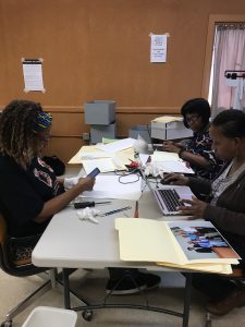 Group of femme-presenting Black people around a table with computers and putting photographs and papers into folders.