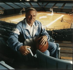 Older white male in Carolina Blue track suit, sitting down, holding a basketball in the UNC arena.