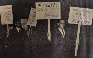 Several white young men in suits carrying signs, some with indiscernible text. Two signs read "New Left: Silent Sponsor" and "Don't give your $$$ to the New Left." 