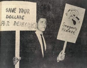 A young white man in a suit and tie holds two signs, one reading "Save your dollars for democracy" and another reading "Seeger's hammer" with a drawing of a hammer and sickle dripping blood.