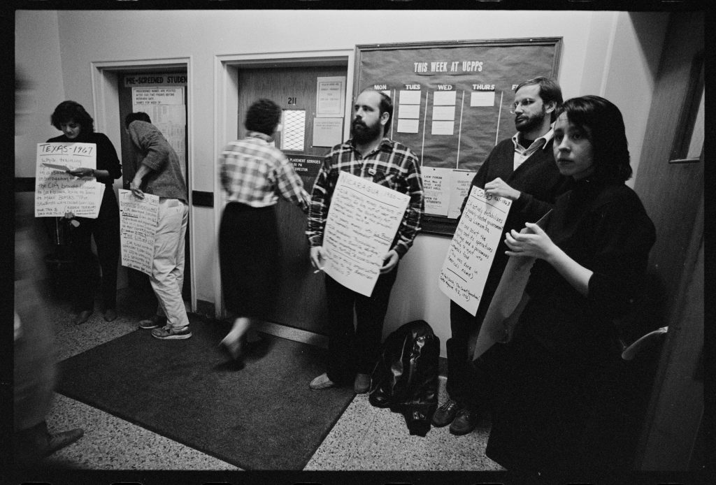 Five students holding signs inside a building on either side of an office door.