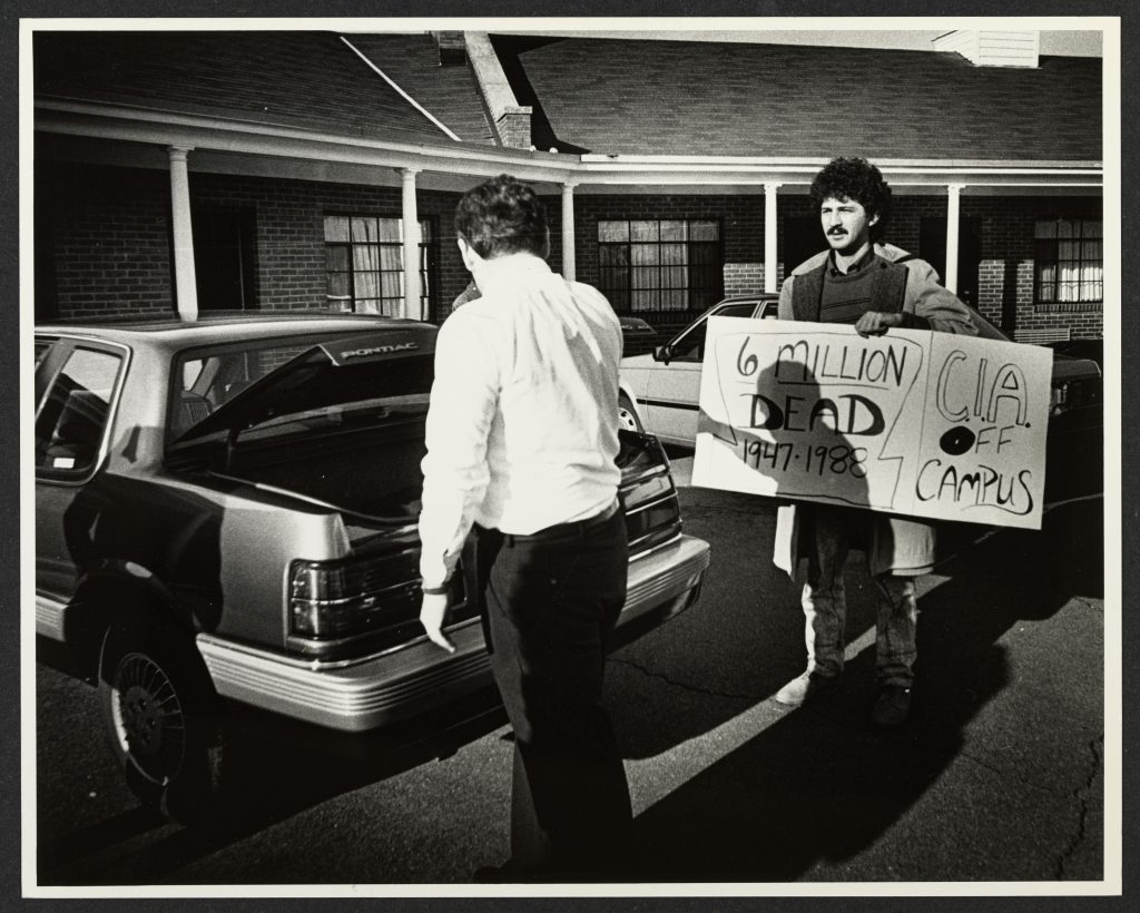 Photo of a student holding a poster reading "6 Million Dead 1947-1988 / CIA Off Campus" while a man places materials in the trunk of a car.