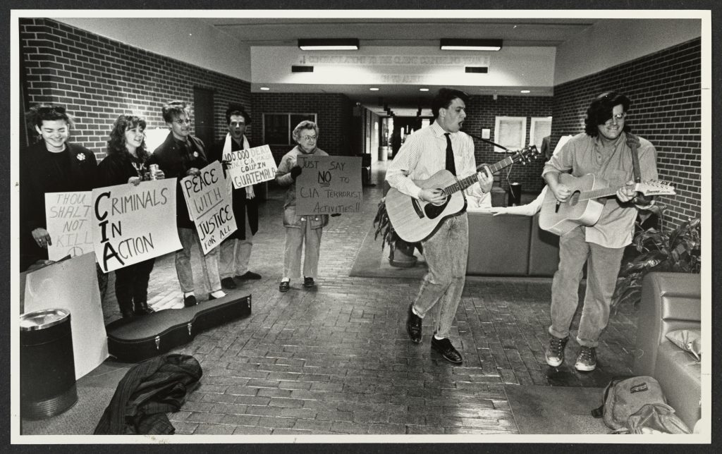 Students protest in a building lobby. Two students are playing acoustic guitars, others are holding signs. The legible signs say "Criminals In Action" and "Peace with Justice for All."